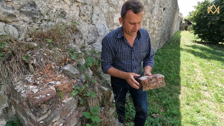 Tomasz Olszacki obok muru trzyma cegłę, moment odkrycia niezwykłego znaleziska